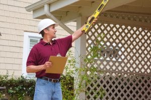 A home inspector or contractor in a hard hat holding a level and a clipboard outside a home doing an inspection or construction quote