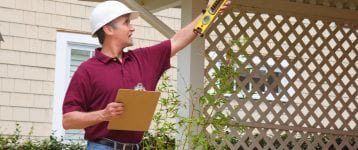 A home inspector or contractor in a hard hat holding a level and a clipboard outside a home doing an inspection or construction quote