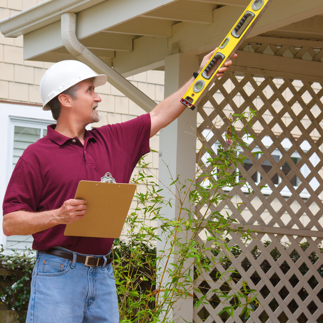 A home inspector or contractor in a hard hat holding a level and a clipboard outside a home doing an inspection or construction quote