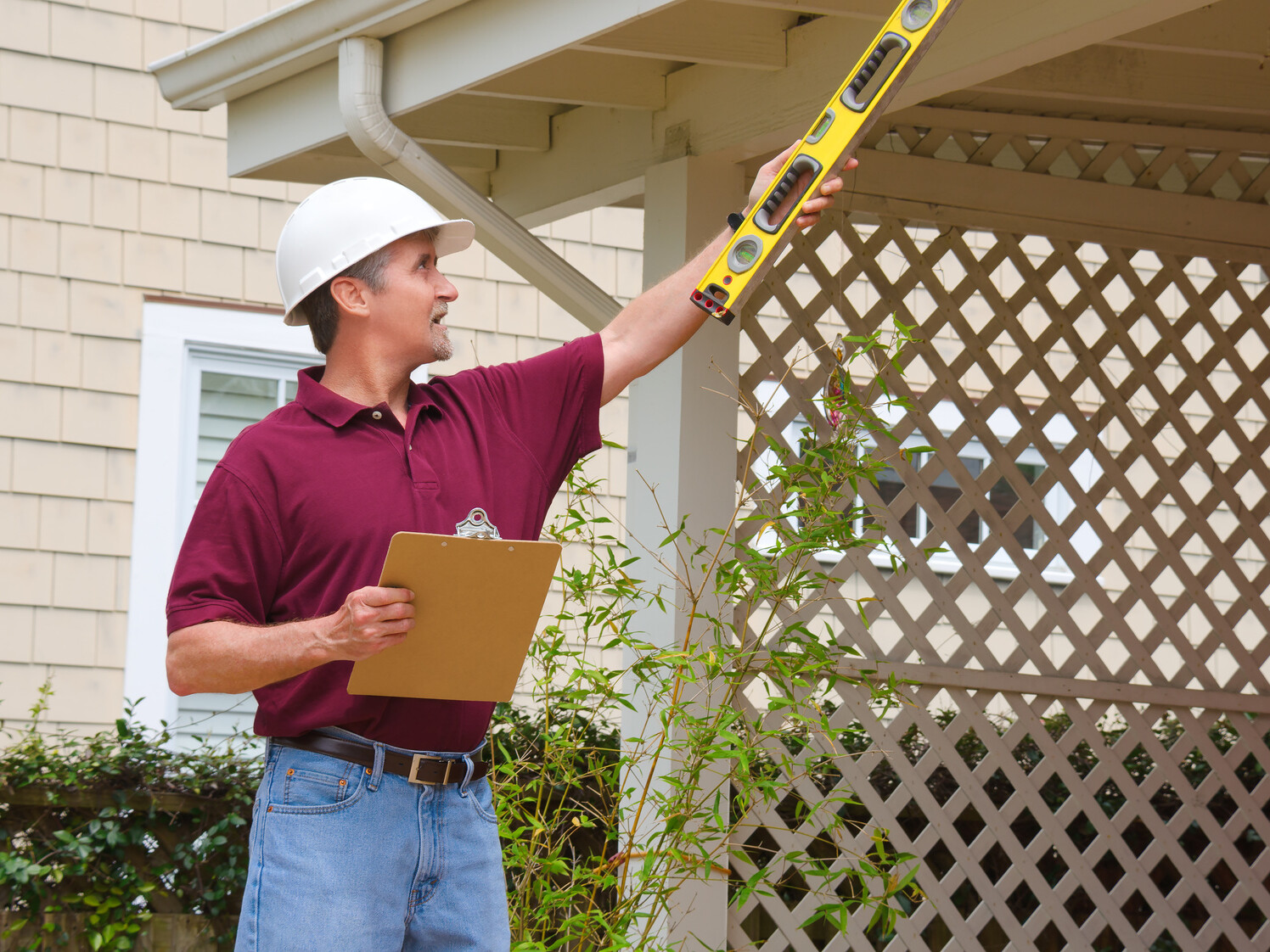 A home inspector or contractor in a hard hat holding a level and a clipboard outside a home doing an inspection or construction quote