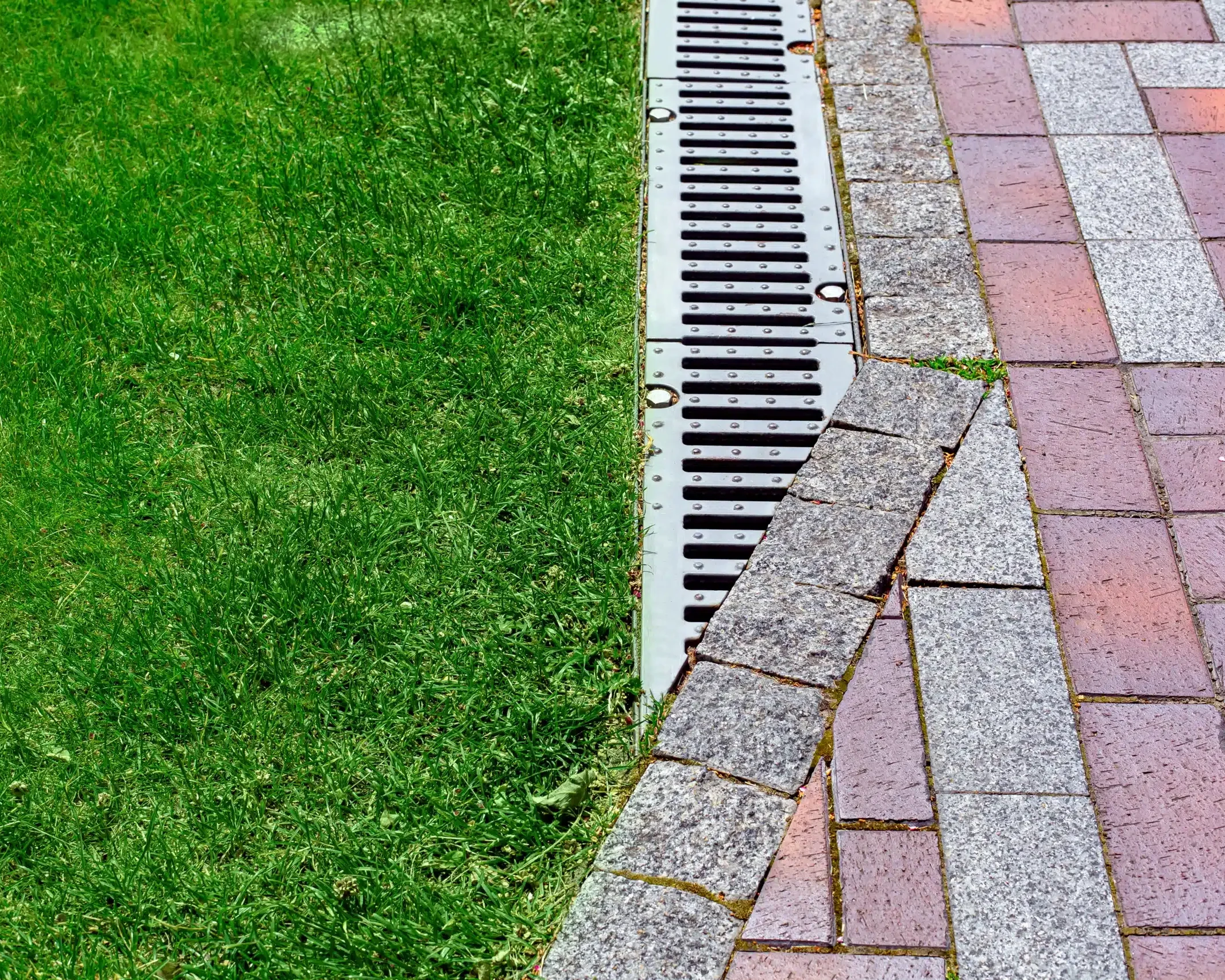 drainage grate bolted to storm drain at corner of pedestrian pavement walk way made of stone brick tiles gray and red pattern in backyard with copy space on green grass on park.