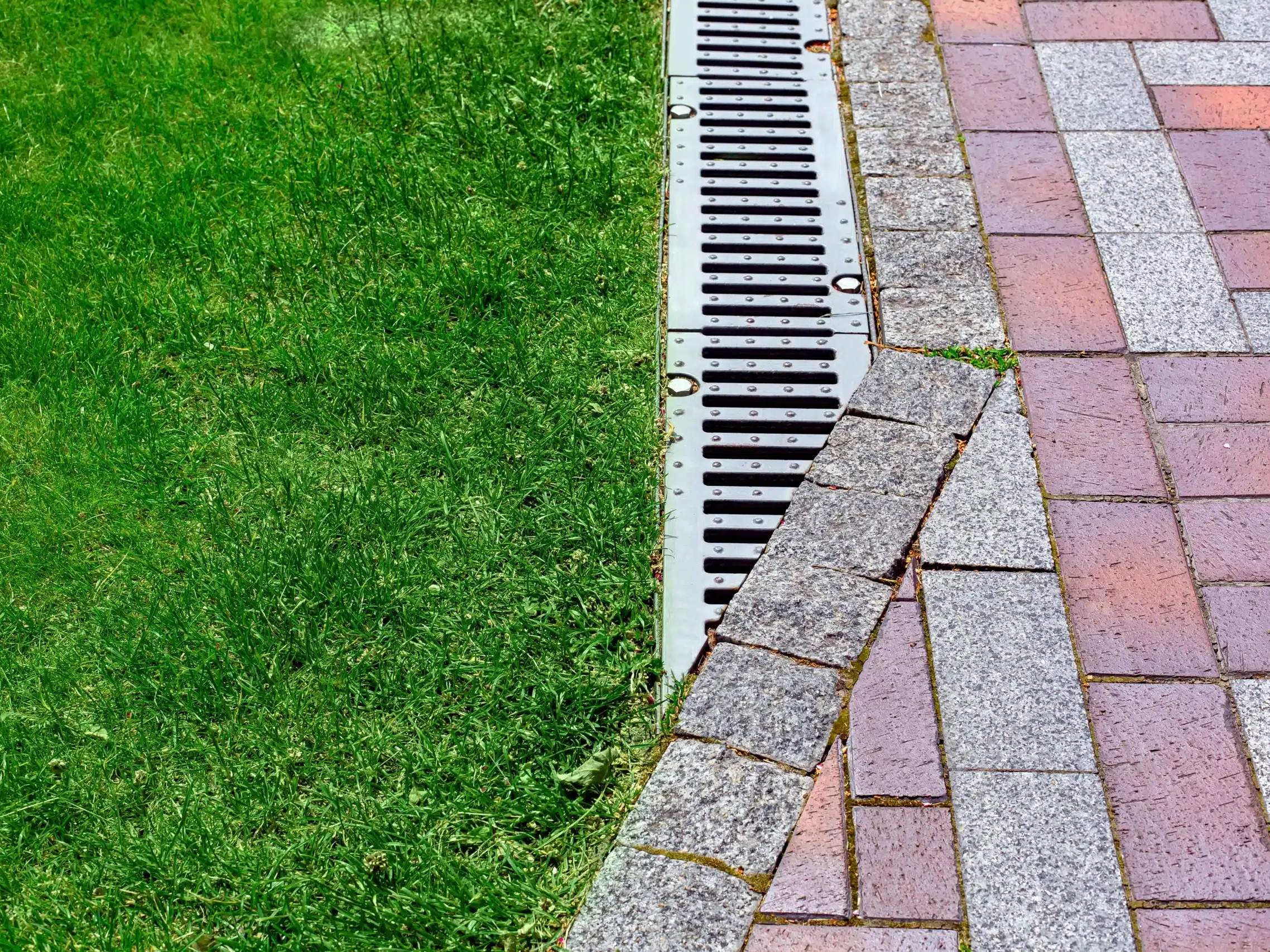 drainage grate bolted to storm drain at corner of pedestrian pavement walk way made of stone brick tiles gray and red pattern in backyard with copy space on green grass on park.