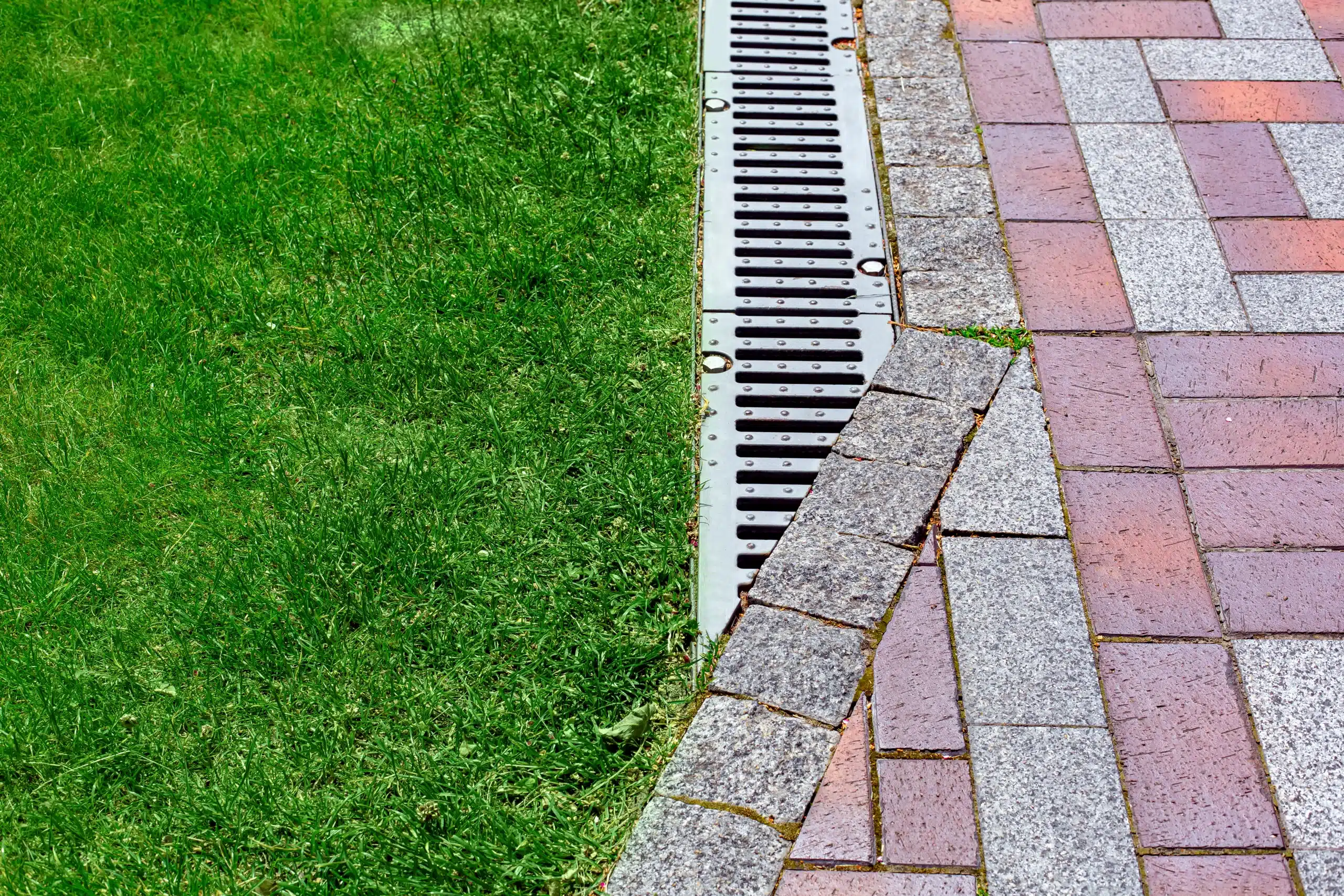 drainage grate bolted to storm drain at corner of pedestrian pavement walk way made of stone brick tiles gray and red pattern in backyard with copy space on green grass on park.