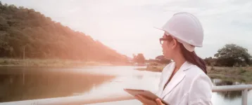 Female engineer in a white helmet working with a digital tablet and looking away at the dam construction site to generate electricity.