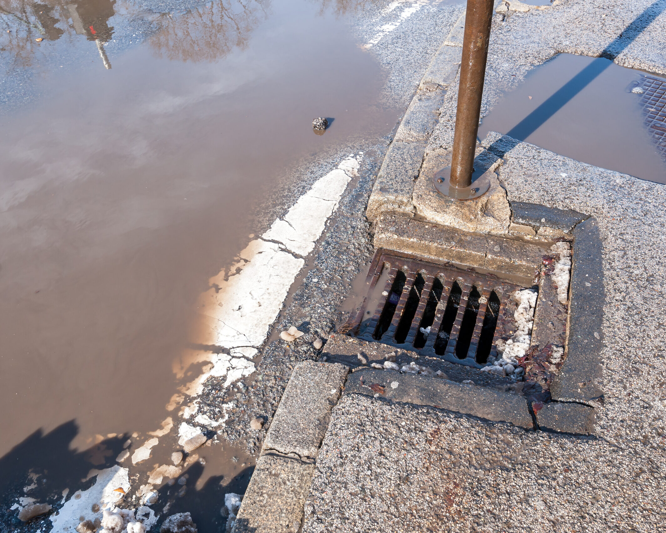 Drainage or sewage manhole grid system on the street asphalt road useless because of damaged tarmac with pot hole full of water after heavy rain and melted snow in the winter season